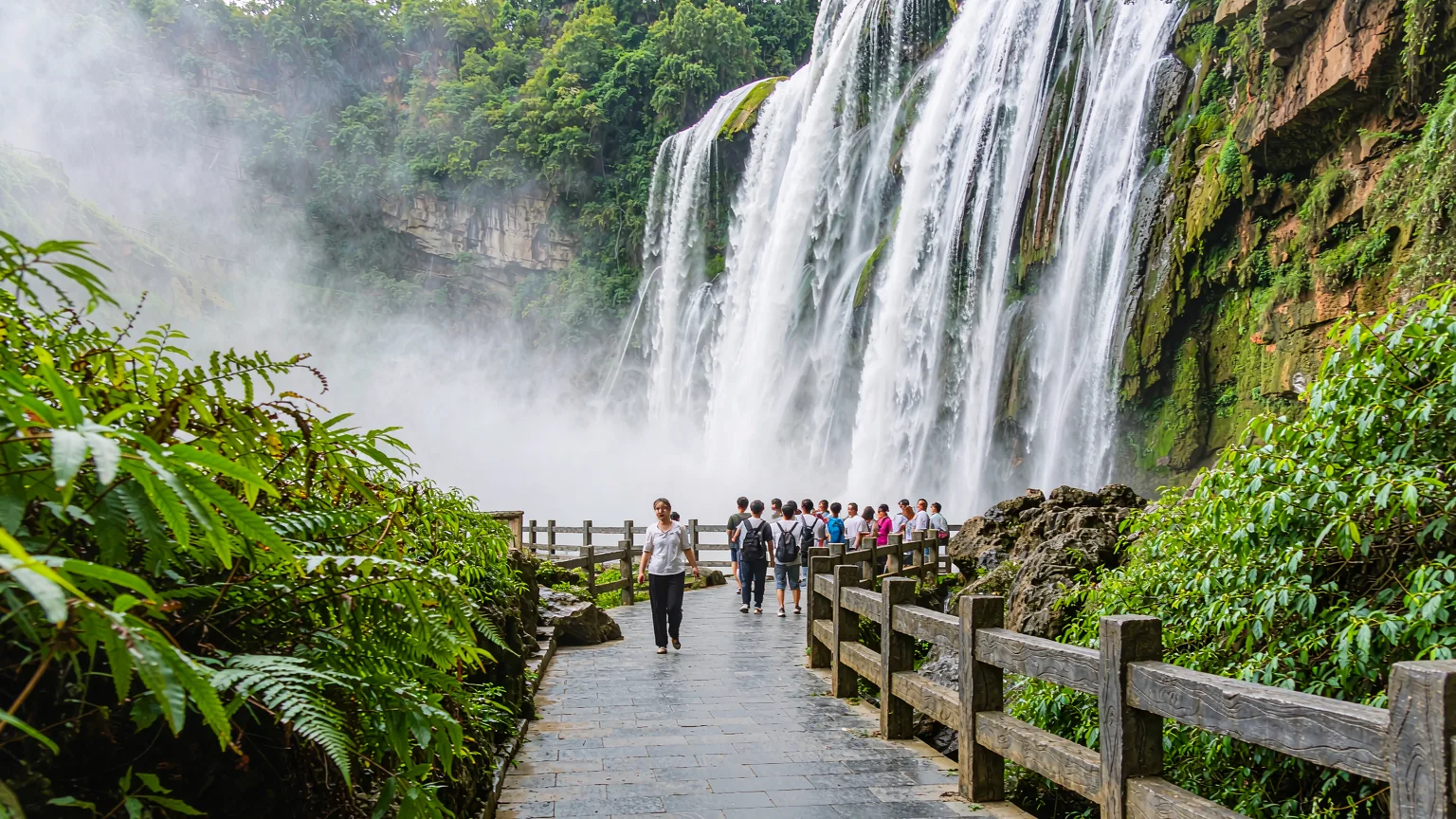 Tourists on stone pathway approaching Huangguoshu Waterfall viewing platform amid mist