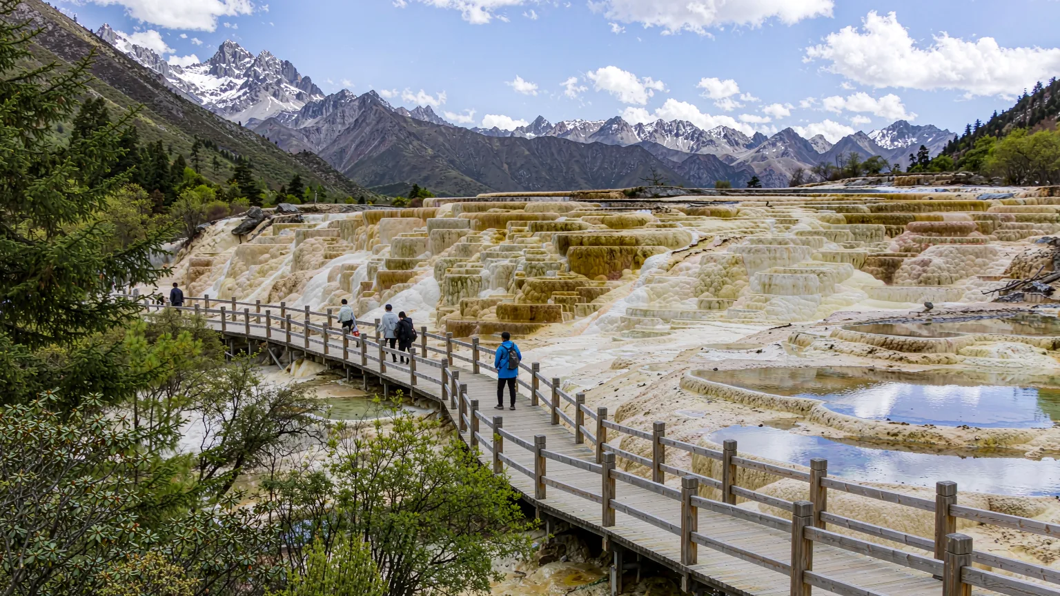 Wooden boardwalk through Huanglong's travertine valley with hikers and mountain views