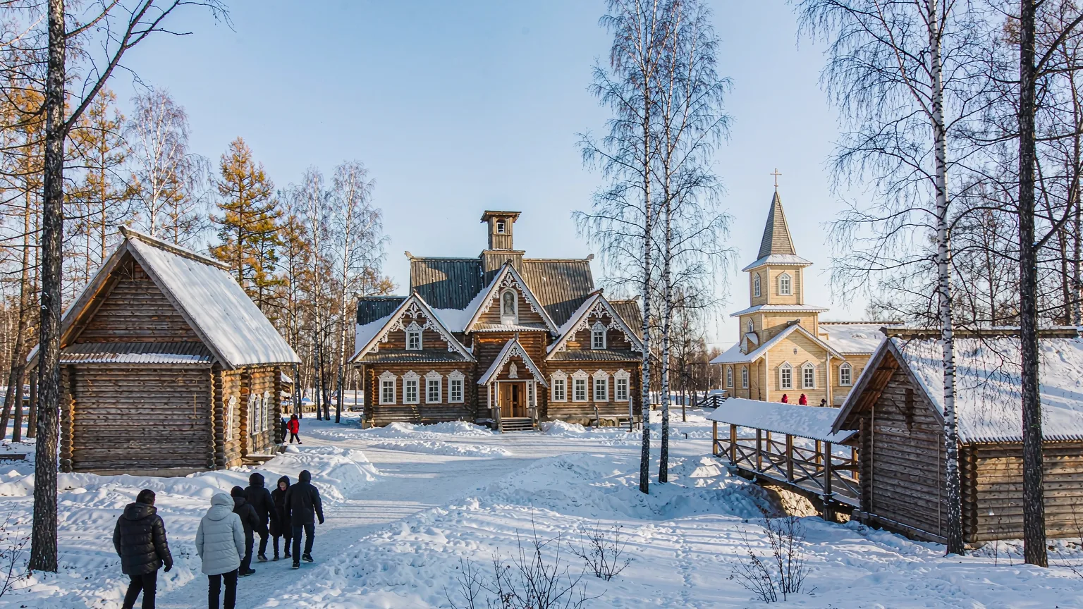 Visitors walking past wooden manor house and church at Volga Manor in winter